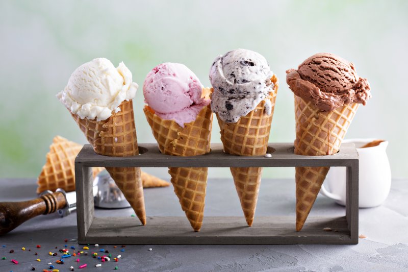 Four ice cream cones sitting in a stand. From left to right: vanilla, strawberry, cookies and cream, chocolate. An ice cream scoop, waffle cone, and some toppings are in the background.